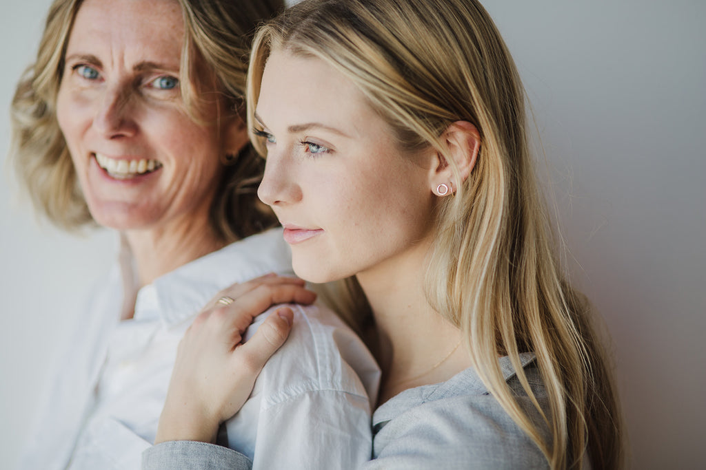 Daughter standing behind her mother. The daughter is wearing the solid gold mini water circle earrings, the solid gold 'Piece of Me' chain, and solid gold stacking rings.  The mother is waring the solid gold medium Water Circle Earrings.